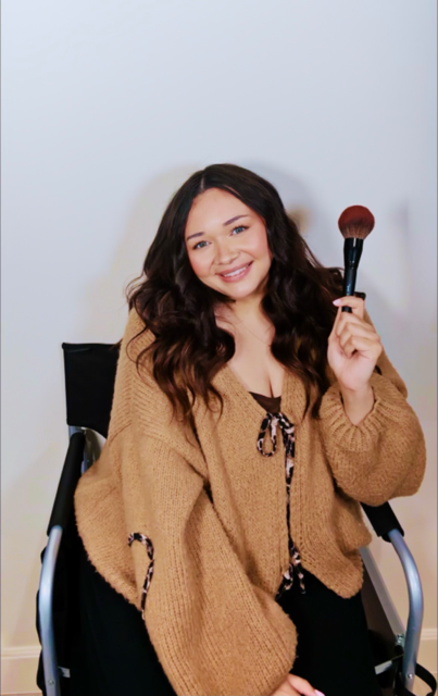 Katelyn Marie holding a makeup brush and smiling in her artist chair.